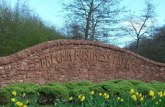 A curved red stone wall featuring the words "Brechin Business Park" carved into the center. Green bushes and yellow daffodils are planted along the base of the wall.