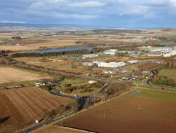 An elevated aerial view of a business park situated among rural farmland. A main road and a roundabout are in the foreground, with several low-rise office buildings and a body of water in the distance.