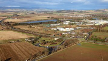An elevated aerial view of a business park situated among rural farmland. A main road and a roundabout are in the foreground, with several low-rise office buildings and a body of water in the distance.