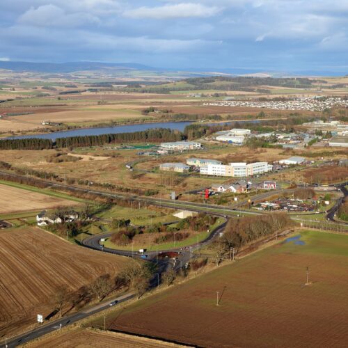 An elevated aerial view of a business park situated among rural farmland. A main road and a roundabout are in the foreground, with several low-rise office buildings and a body of water in the distance.