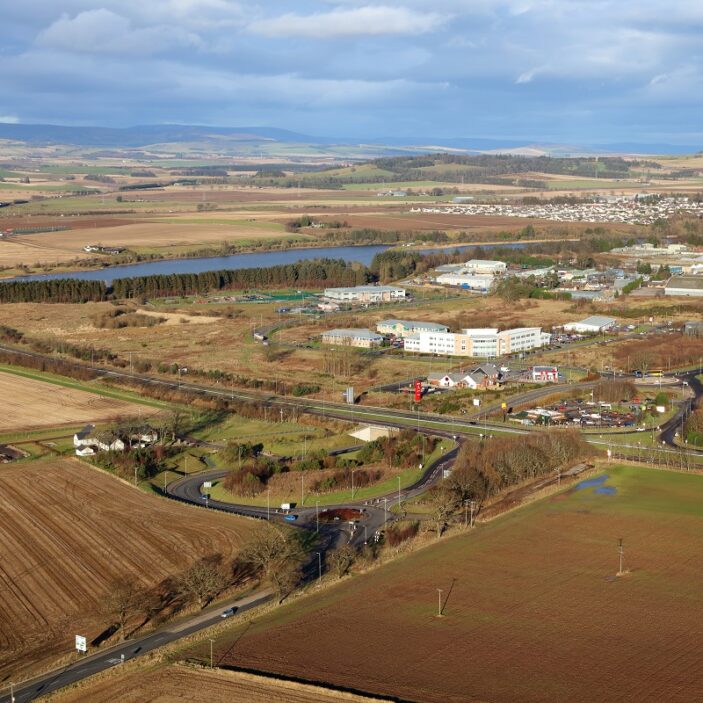 An elevated aerial view of a business park situated among rural farmland. A main road and a roundabout are in the foreground, with several low-rise office buildings and a body of water in the distance.
