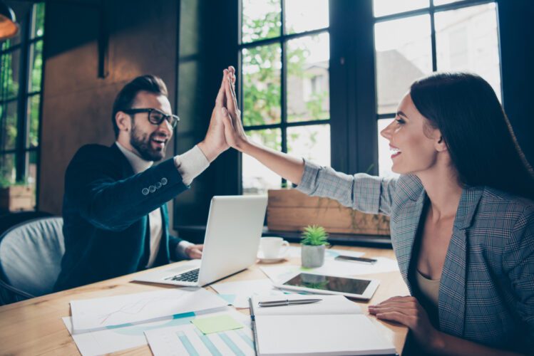 A man and a woman in professional attire share an enthusiastic high-five across a wooden office desk. Both are smiling broadly in a bright, modern workspace filled with natural light from large windows. The desk is scattered with a laptop, tablet, notebooks, and business charts.