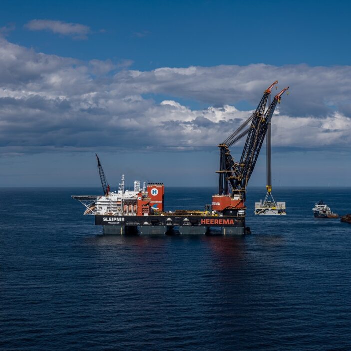 The semi-submersible crane vessel "Sleipnir" uses its massive dual cranes to lower a module onto a yellow offshore foundation in the middle of a calm blue ocean.