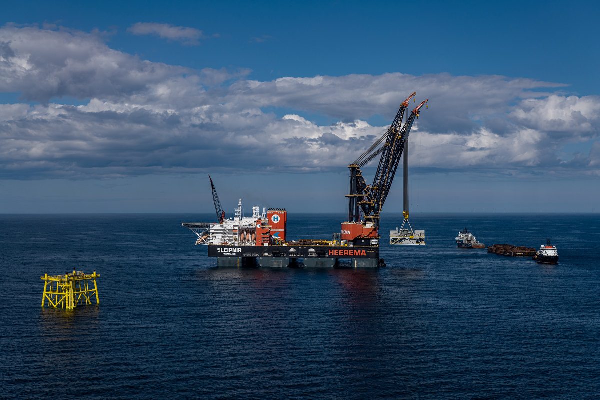 The semi-submersible crane vessel "Sleipnir" uses its massive dual cranes to lower a module onto a yellow offshore foundation in the middle of a calm blue ocean.
