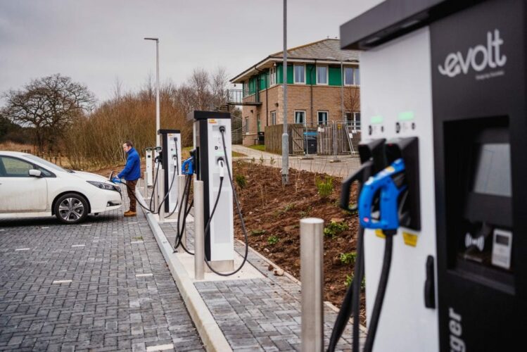 A man in a blue jacket plugs a charging cable into a white electric car at a row of Evolt rapid charging stations in a paved parking area at Orchardbank Business Park.