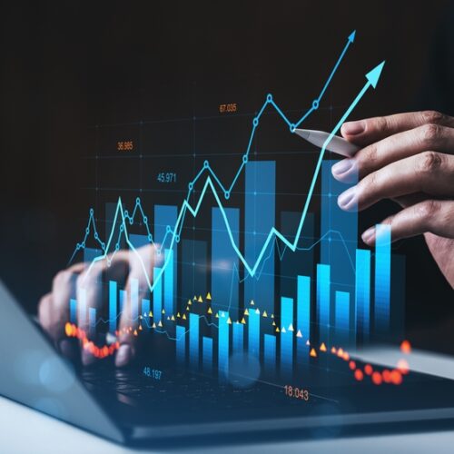 A person uses a stylus to interact with a glowing, 3D holographic bar chart and line graph rising from a laptop keyboard. Blue arrows and data points indicate an upward trend, while a smaller orange dotted line shows a lower trajectory against a dark background.