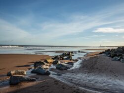 A wide-angle landscape of Montrose Beach at low tide, featuring a line of large rocks cutting through wet, footprint-marked sand leading toward the ocean. A small stream of water reflects the bright blue sky, which is filled with soft, wispy white clouds. In the far distance, a thin white lighthouse sits on the horizon under a clear, expansive sky.