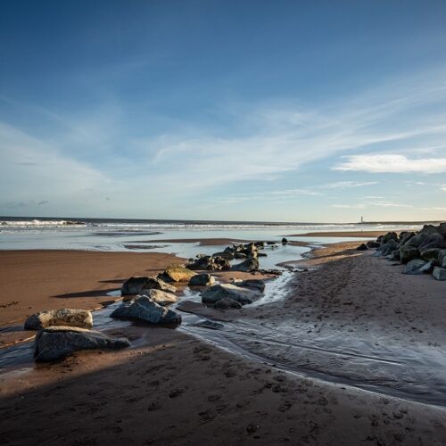 A wide-angle landscape of Montrose Beach at low tide, featuring a line of large rocks cutting through wet, footprint-marked sand leading toward the ocean. A small stream of water reflects the bright blue sky, which is filled with soft, wispy white clouds. In the far distance, a thin white lighthouse sits on the horizon under a clear, expansive sky.