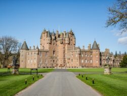 A grand, pink-walled Scottish castle with numerous turrets, spires, and a central clock tower stands under a bright blue sky. A long, paved driveway leads directly to the entrance, flanked by vast green lawns and stone pedestals topped with statues.