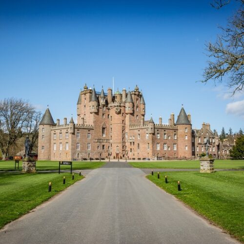 A grand, pink-walled Scottish castle with numerous turrets, spires, and a central clock tower stands under a bright blue sky. A long, paved driveway leads directly to the entrance, flanked by vast green lawns and stone pedestals topped with statues.