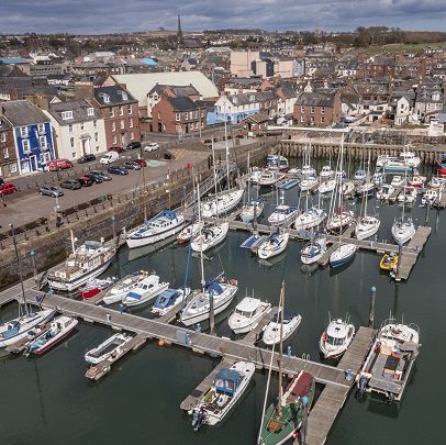 An elevated view of Arbroath Harbour filled with numerous white sailboats and small motorboats moored at wooden piers. The background features a coastal town with stone and brick buildings, a parking area with several cars, and distant church spires under a cloudy sky.