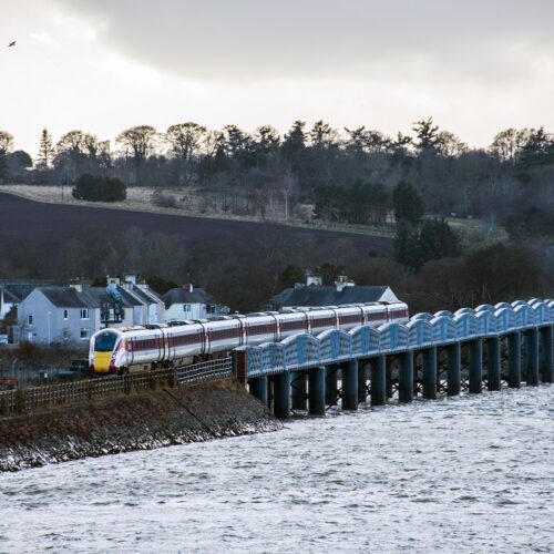 A long white and red passenger train crosses a blue metal railway viaduct over a body of water toward a small town. In the background, rolling hills, dense trees, and a stone church tower are visible under a bright, overcast sky.