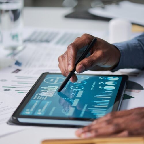 A close-up shot of a person using a stylus to interact with complex data visualizations and charts on a digital tablet, surrounded by printed business reports and a coffee mug on a desk.