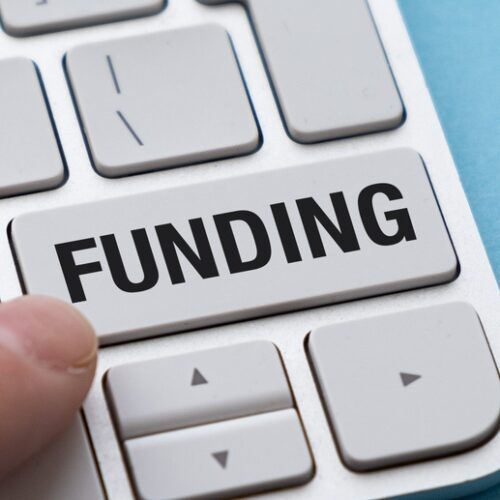 A close-up, top-down view of a finger pressing a large, white computer key labeled "FUNDING" on a modern silver keyboard against a light blue background.