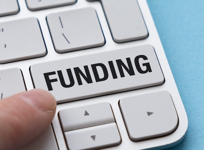 A close-up, top-down view of a finger pressing a large, white computer key labeled "FUNDING" on a modern silver keyboard against a light blue background.
