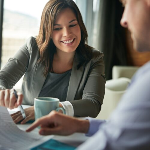 A smiling businesswoman in a grey blazer sits at a desk, reviewing and pointing at a document during a professional meeting with a colleague.