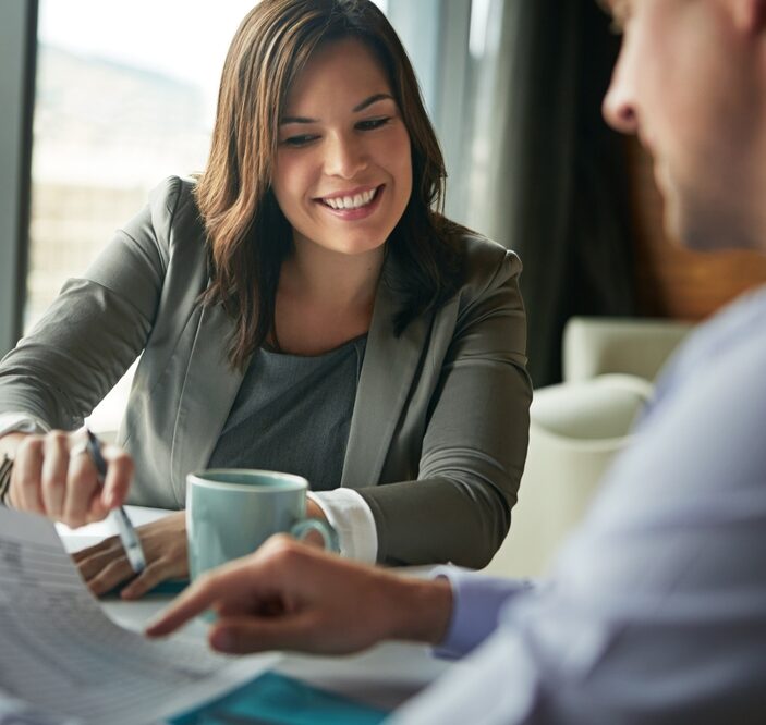 A smiling businesswoman in a grey blazer sits at a desk, reviewing and pointing at a document during a professional meeting with a colleague.