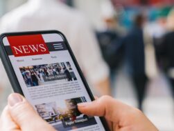 A close-up of a person's hands holding a smartphone displaying a digital news application with headlines like "Public Opinion" and "Public Transport," set against a blurred background of people walking in a busy public space.