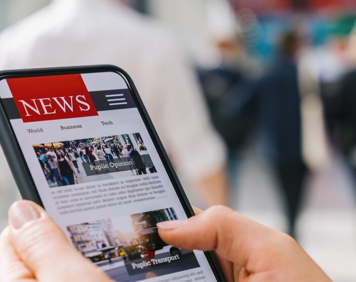 A close-up of a person's hands holding a smartphone displaying a digital news application with headlines like "Public Opinion" and "Public Transport," set against a blurred background of people walking in a busy public space.