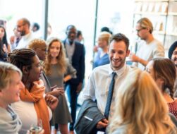 A bright, candid shot of a diverse group of professionals networking and laughing during a daytime event in a modern, sunlit indoor space.