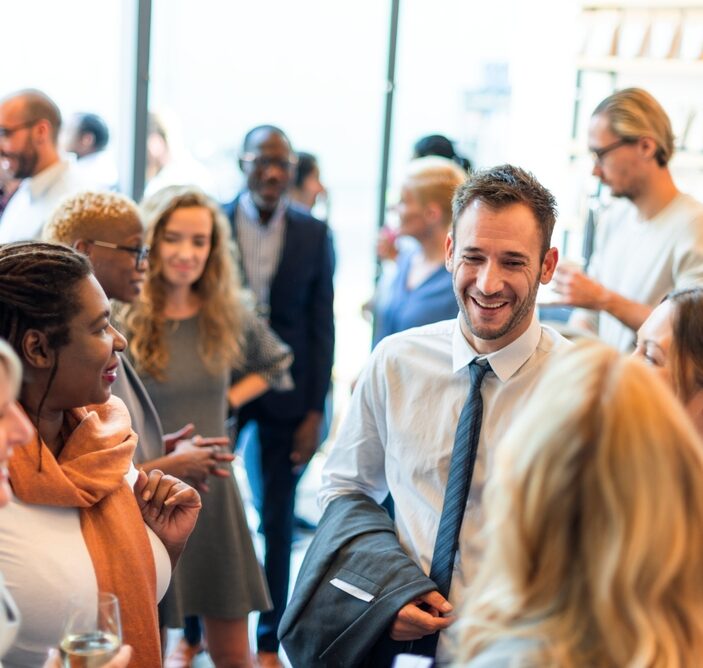 A bright, candid shot of a diverse group of professionals networking and laughing during a daytime event in a modern, sunlit indoor space.