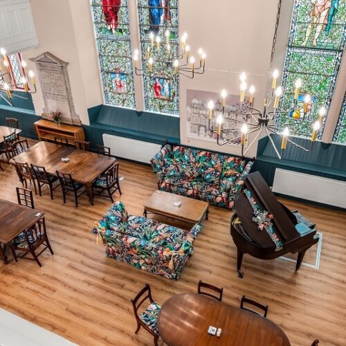 An elevated view shows a spacious community hall or converted church interior with high ceilings and large stained-glass windows. The room is furnished with several dark wood dining tables, a grand piano, and vibrant floral-patterned sofas. Three large, modern chandeliers illuminate the space, which features light wood flooring and deep teal accents along the lower walls.