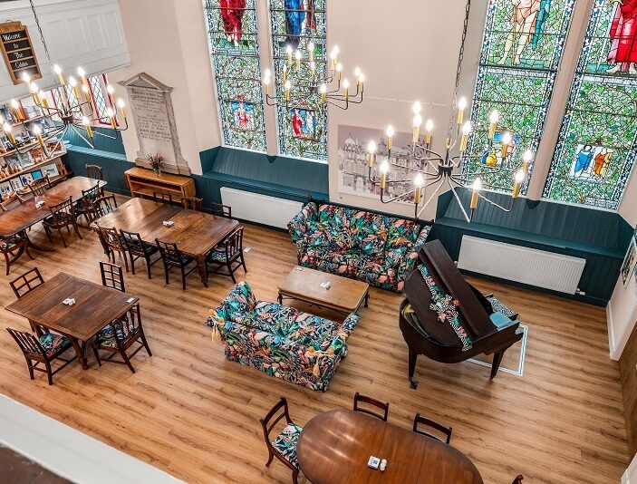 An elevated view shows a spacious community hall or converted church interior with high ceilings and large stained-glass windows. The room is furnished with several dark wood dining tables, a grand piano, and vibrant floral-patterned sofas. Three large, modern chandeliers illuminate the space, which features light wood flooring and deep teal accents along the lower walls.
