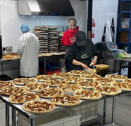 Three staff members in a commercial kitchen work at stainless steel stations to prepare dozens of pizzas. The foreground features a large table crowded with rows of topped pizzas, while the background shows a person in a white lab coat and hairnet near an oven, another in a red hoodie near a tall stack of pizza crusts, and a third in a black apron preparing ingredients.