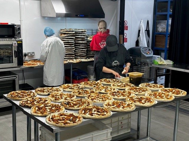 Three staff members in a commercial kitchen work at stainless steel stations to prepare dozens of pizzas. The foreground features a large table crowded with rows of topped pizzas, while the background shows a person in a white lab coat and hairnet near an oven, another in a red hoodie near a tall stack of pizza crusts, and a third in a black apron preparing ingredients.