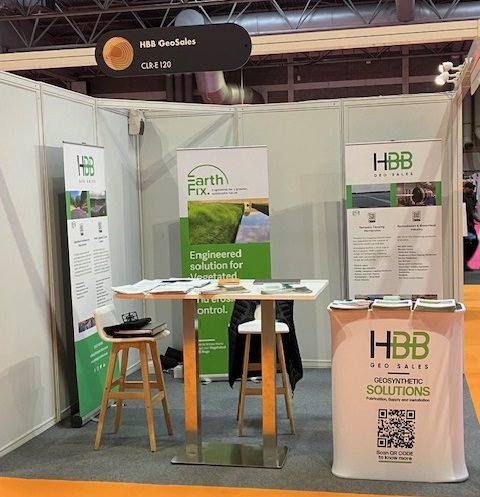 An indoor exhibition booth for HBB GeoSales features three tall roll-up banners and a branded reception counter. The displays highlight "Geosynthetic Solutions" and "Earth Fix" engineered solutions for erosion control, using green and white branding. A high-top table with two stools sits in the center of the booth, which is positioned on a grey carpeted area within a larger event hall.