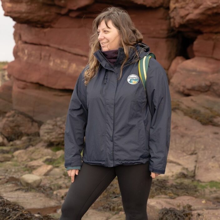 A woman stands on a rocky shore in front of a massive, layered red sandstone cliff. She is wearing a dark navy waterproof jacket with a small circular logo and black leggings, looking off to the side with a slight smile. The ground around her is covered in dark seaweed and scattered stones.