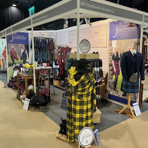 An exhibition stand for Strathmore Woollen Co. showcasing various traditional Scottish textiles. The booth features mannequins dressed in kilts and jackets, alongside racks and displays of tartan fabrics, including a prominent yellow and black tartan draped in the foreground. Large posters on the walls display models wearing the company's woollen clothing.