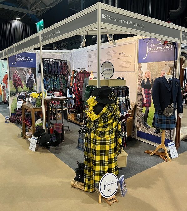 An exhibition stand for Strathmore Woollen Co. showcasing various traditional Scottish textiles. The booth features mannequins dressed in kilts and jackets, alongside racks and displays of tartan fabrics, including a prominent yellow and black tartan draped in the foreground. Large posters on the walls display models wearing the company's woollen clothing.