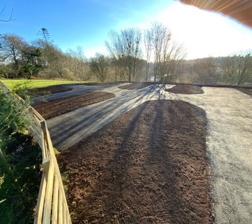 An outdoor construction site shows a newly paved asphalt driveway or parking area surrounded by fresh dark brown soil beds. A wooden fence runs along the left side, and several leafless trees are visible in the background under a bright, clear blue sky with the sun shining directly into the frame.
