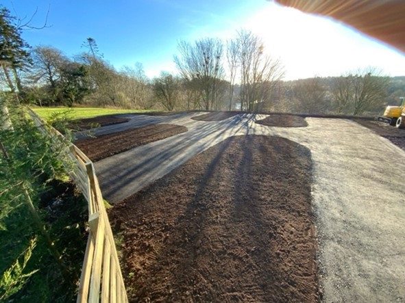 An outdoor construction site shows a newly paved asphalt driveway or parking area surrounded by fresh dark brown soil beds. A wooden fence runs along the left side, and several leafless trees are visible in the background under a bright, clear blue sky with the sun shining directly into the frame.