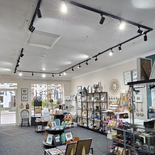 Gemini said The interior of a brightly lit gift shop features black track lighting on the ceiling illuminating various displays of home goods and art. Shelves and tables are filled with items such as ceramics, framed pictures, candles, and stationary. A large glass storefront window and a door at the back look out onto a street, allowing natural light to enter the space.