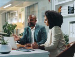 Two business professionals, a man in a green suit and a woman in a beige blazer, sit at a white desk in a modern office, collaborating while looking at a laptop screen together.