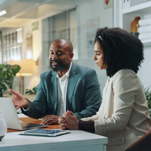Two business professionals, a man in a green suit and a woman in a beige blazer, sit at a white desk in a modern office, collaborating while looking at a laptop screen together.
