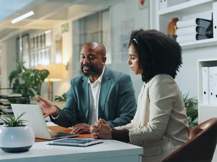 Two business professionals, a man in a green suit and a woman in a beige blazer, sit at a white desk in a modern office, collaborating while looking at a laptop screen together.