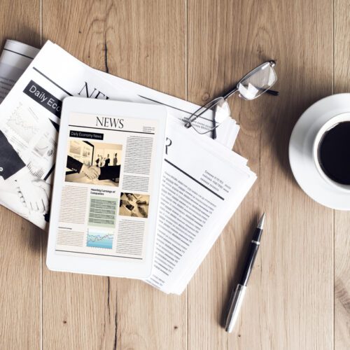 A top-down view of a wooden desk featuring a tablet displaying a digital news site resting on top of a physical newspaper, accompanied by a cup of black coffee, a pair of glasses, and a silver pen.