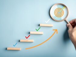 A conceptual image featuring a staircase of wooden blocks with colorful check marks above them, leading toward a target revealed through a magnifying glass held by a hand.