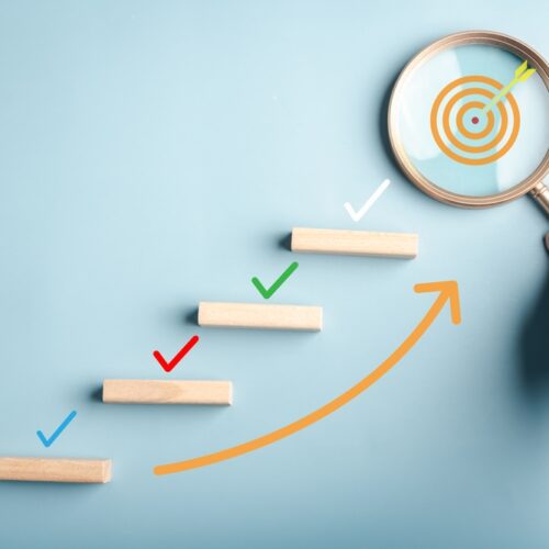 A conceptual image featuring a staircase of wooden blocks with colorful check marks above them, leading toward a target revealed through a magnifying glass held by a hand.