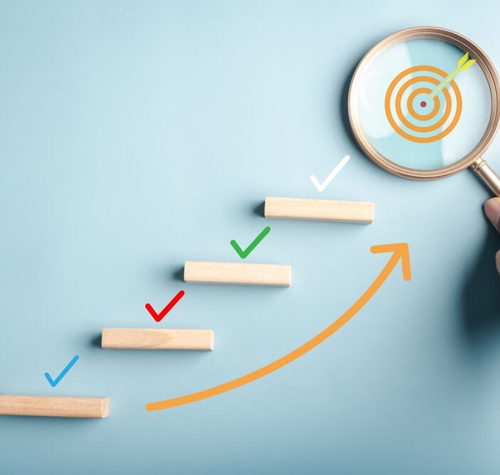 A conceptual image featuring a staircase of wooden blocks with colorful check marks above them, leading toward a target revealed through a magnifying glass held by a hand.