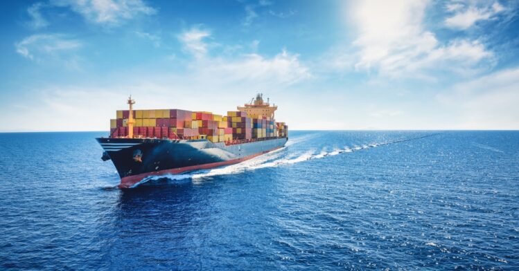 A large container ship loaded with multicolored shipping containers cruises through a calm, bright blue ocean under a sunny sky with scattered clouds.