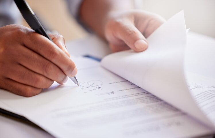 A close-up of a person's hands as they sign a formal paper document with a silver pen. One hand holds the pen to write a signature, while the other holds back the corner of the page.