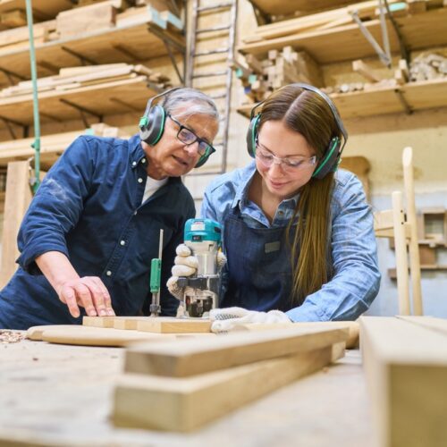 Two carpenters working in a woodworking shop; a woman uses a power router on a piece of wood while an older man looks on and assists. Both are wearing safety gear, including ear protection and glasses, surrounded by lumber and workshop tools.