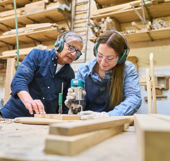 Two carpenters working in a woodworking shop; a woman uses a power router on a piece of wood while an older man looks on and assists. Both are wearing safety gear, including ear protection and glasses, surrounded by lumber and workshop tools.
