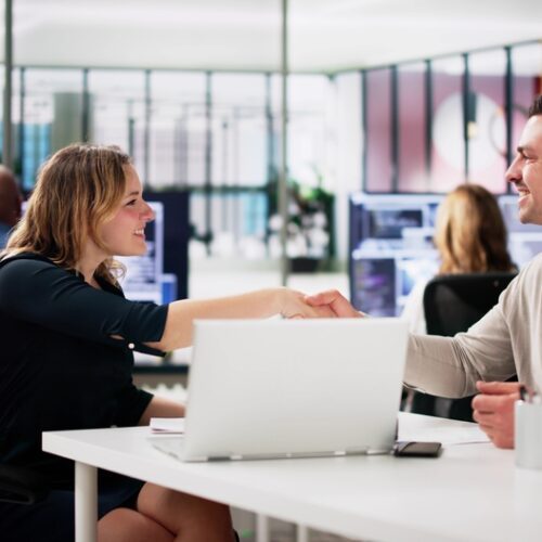 A man and woman sitting at a desk in a modern, open-plan office, shaking hands and smiling at each other. A laptop is visible on the white desk between them, and other colleagues are blurred in the background.