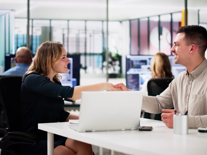 A man and woman sitting at a desk in a modern, open-plan office, shaking hands and smiling at each other. A laptop is visible on the white desk between them, and other colleagues are blurred in the background.