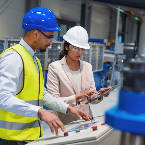 An engineer and a technician in a factory setting, both wearing hard hats and safety glasses. The engineer uses a tablet while the technician operates a control panel on a piece of large blue industrial machinery.